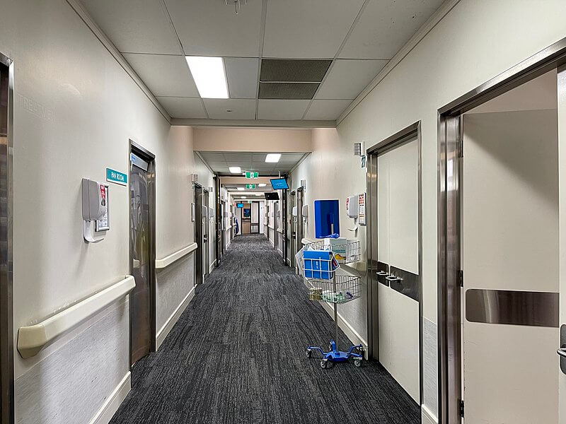A long, well-lit hospital hallway with patient rooms on both sides, featuring metal doors and signage. Medical equipment, including a supply cart, is placed along the walls, and the floor is covered with dark carpet.
