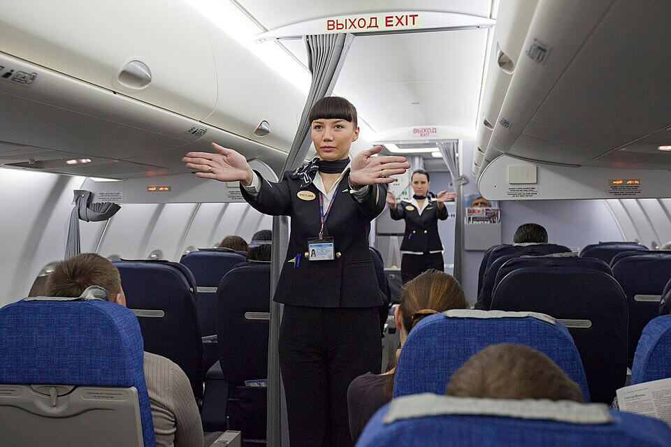 Two flight attendants in dark uniforms stand in the aisle of an airplane, demonstrating safety procedures to passengers. With serious expressions, they extend their arms to illustrate emergency exits, while seated travelers watch attentively or look down.