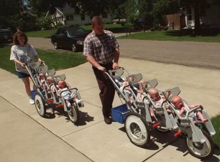 A man and a woman push two large multi-seat strollers filled with several babies down a sunny suburban sidewalk. The babies, dressed in light-colored outfits, are securely strapped in, some appearing content while others squirm in their seats.
