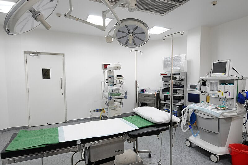 A brightly lit, sterile operating room with white walls and a gray floor. The room contains an operating table with a green and white surgical drape, overhead surgical lights, medical monitors, an anesthesia machine, and various medical equipment neatly arranged on shelves and carts.