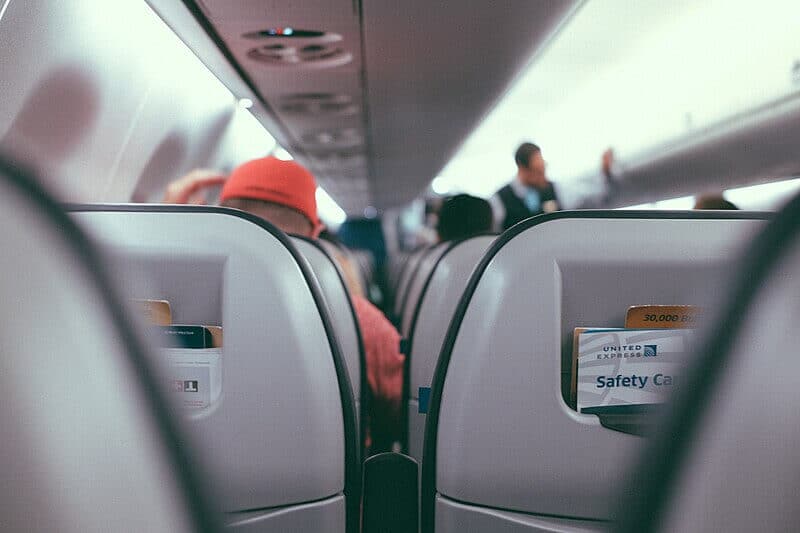 A view from the middle of an airplane cabin shows rows of seats with safety cards tucked into the pockets. Passengers, including one wearing a red cap, sit and settle in while a flight attendant moves down the aisle.