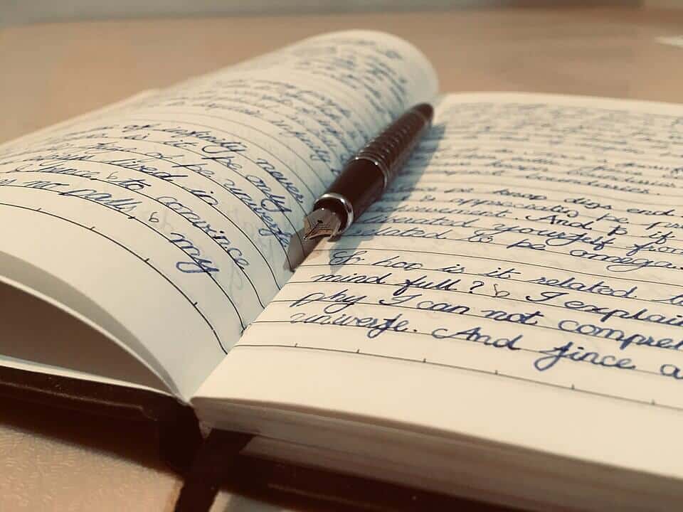 Open notebook filled with neat, cursive handwriting in blue ink, with a fountain pen resting across the pages. The scene is warmly lit, and the focus highlights the pen's metallic nib and the texture of the paper.