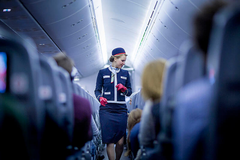 A flight attendant in a navy blue uniform with red gloves walks down the aisle of an airplane, smiling as she interacts with passengers. The dim cabin lighting and rows of seats filled with travelers create a cozy yet bustling in-flight atmosphere.