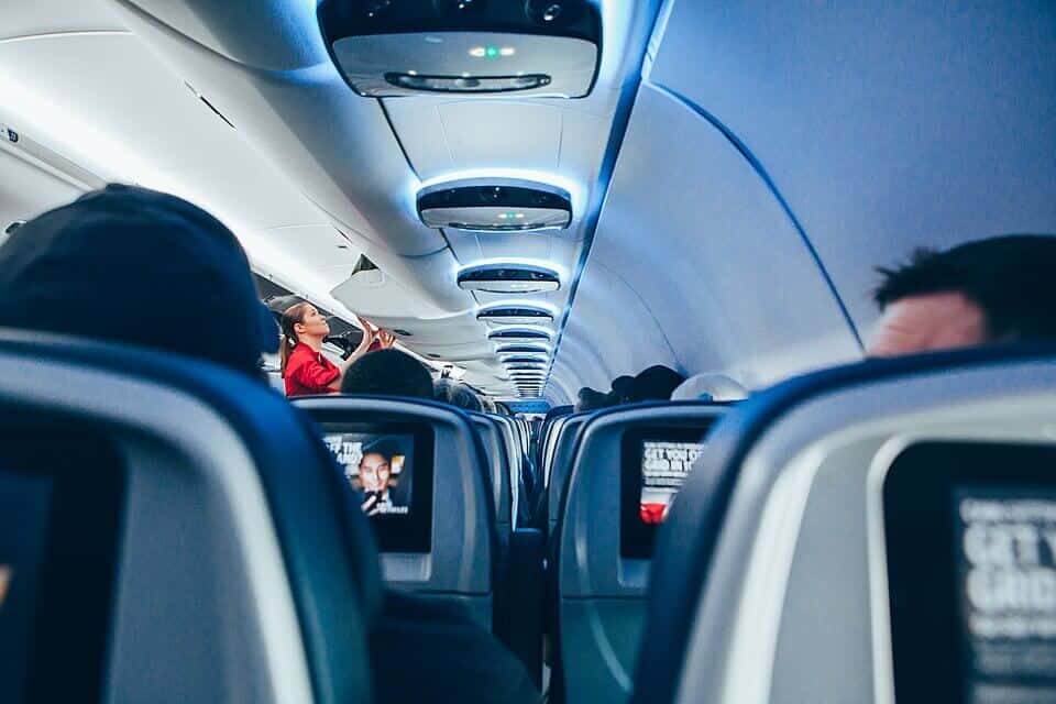 A modern airplane cabin with dim blue lighting shows passengers seated, some watching in-flight entertainment. A flight attendant in a red uniform reaches up to adjust an overhead compartment while travelers settle in for the journey.