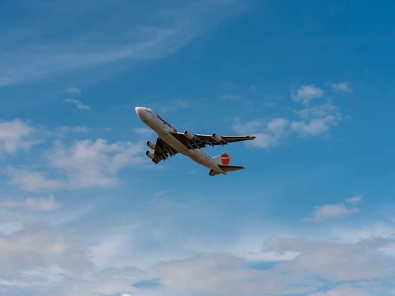 A large commercial airplane soars through a bright blue sky, its engines roaring beneath the wings. Wisps of white clouds scatter across the background as the aircraft ascends gracefully into the open air.