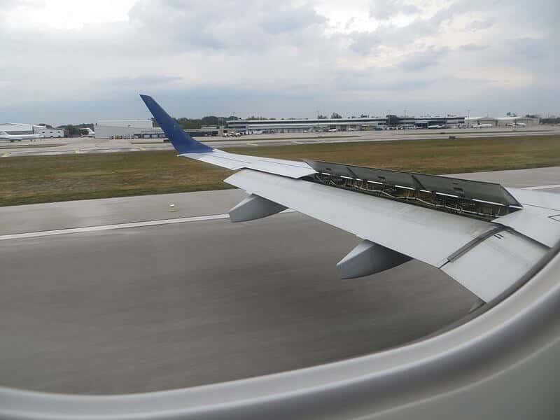 An airplane wing is seen from the passenger window, with its speed brakes deployed as the aircraft lands on the runway. The airport buildings and grassy surroundings stretch into the distance under a cloudy sky, signaling the end of a journey.