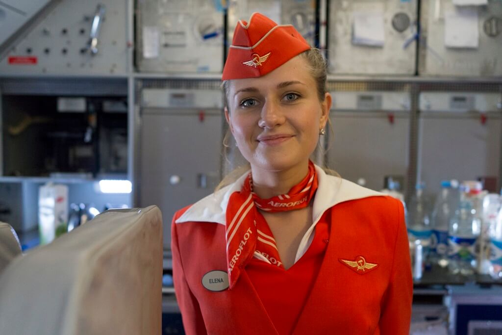 A flight attendant in a bright red uniform and matching hat stands in the airplane galley, smiling warmly. Behind her, stainless steel compartments and beverage supplies line the workspace, creating a busy yet organized atmosphere.