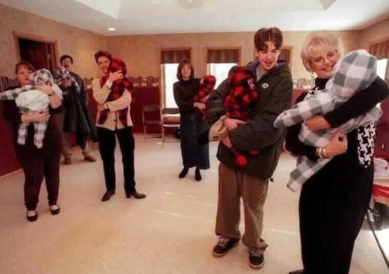 A group of people stand in a spacious room, each holding a baby dressed in either red-and-black or gray-and-white plaid outfits. The individuals, appearing to be a mix of family members and caregivers, are smiling and interacting with the infants.