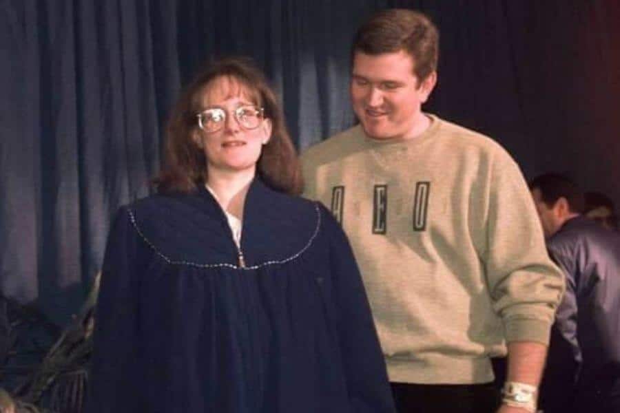 A woman wearing glasses and a dark blue oversized gown walks forward with a neutral expression, while a man in a beige "NED" sweatshirt smiles behind her. He wears a hospital bracelet on his wrist, and the background features a dark curtain with other people present.