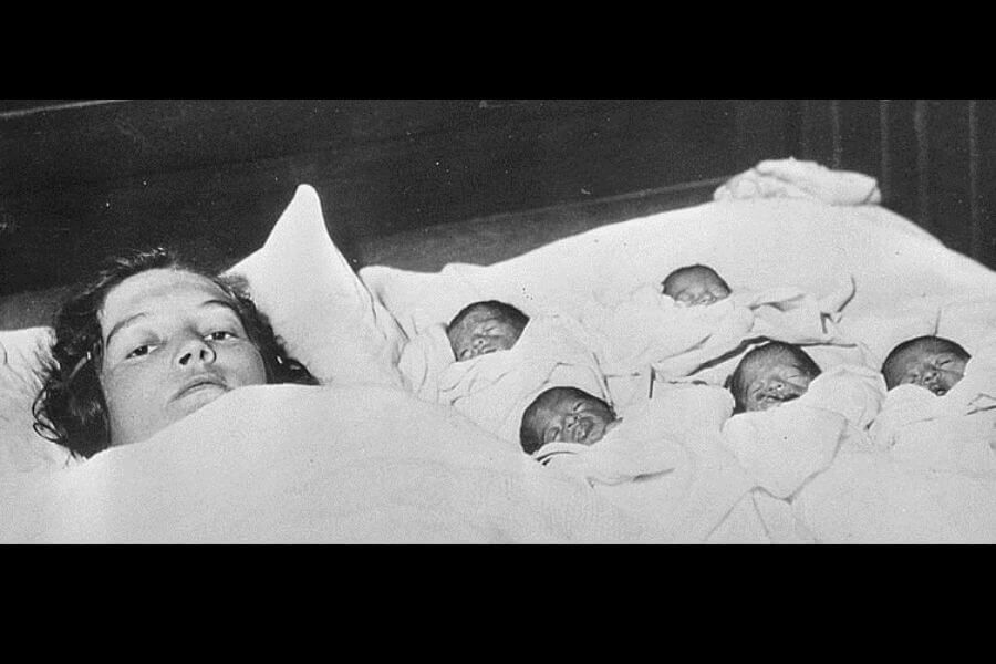 A black-and-white photograph of a mother lying in bed, looking exhausted, with five newborn babies swaddled in blankets beside her. The babies are lined up in a row, sleeping peacefully, while the mother rests her head on a pillow.