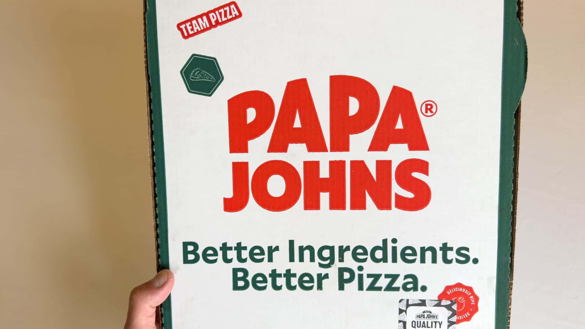 San Jose, CA - June 1, 2023: Hand holding a Papa Johns pizza cardboard box against tan background.
