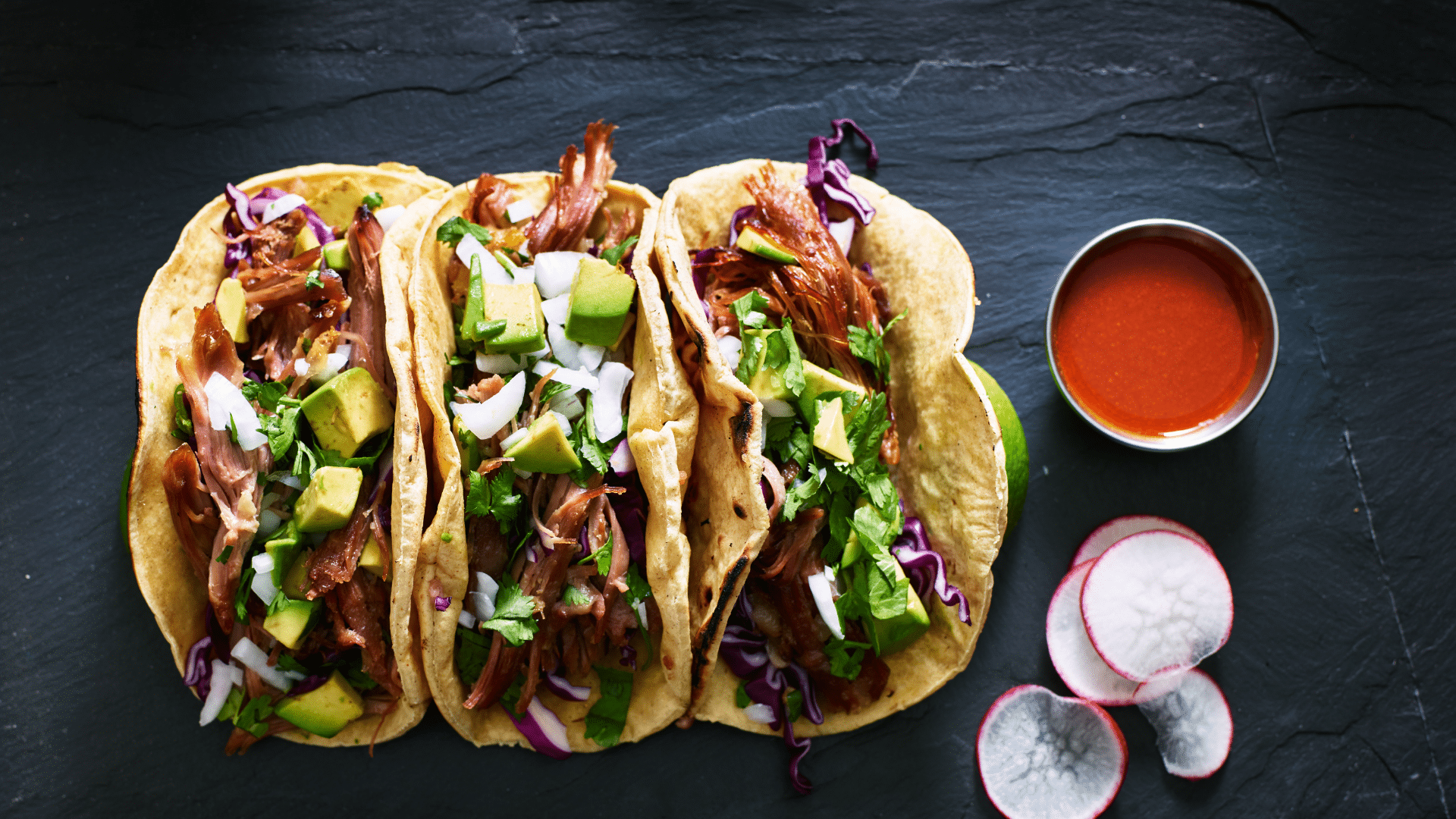 Three pork carnitas tacos served with red hot sauce on a flatbread