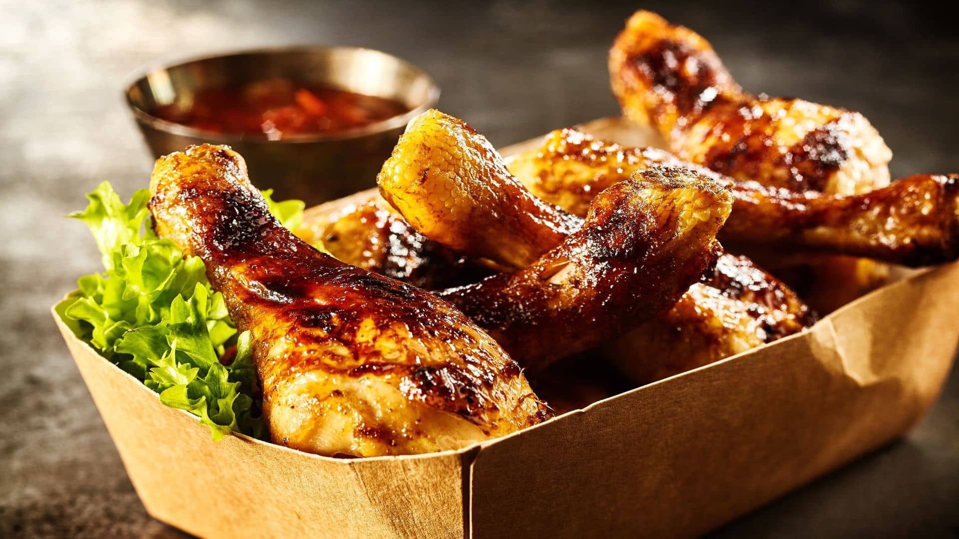 Takeaway box of crispy grilled chicken legs with a spicy marinade served with fresh lettuce and a hot chili dipping sauce, close up view