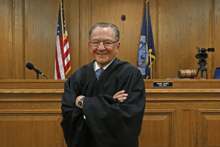 Judge Frank Caprio stands confidently in his courtroom, arms folded, with flags and a camera setup behind him.