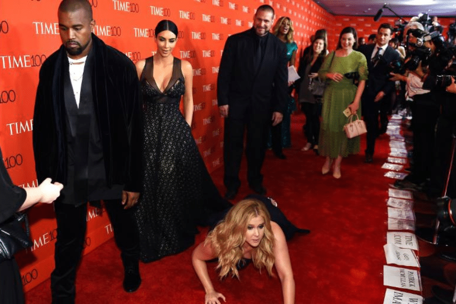 Celebrities on a red carpet; one person dramatically pretends to fall, creating a humorous moment. Onlookers and photographers capture the scene.