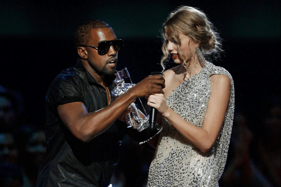 Man in sunglasses interrupting a woman in a sparkling dress on stage, holding a microphone and trophy. The atmosphere is tense and unexpected.