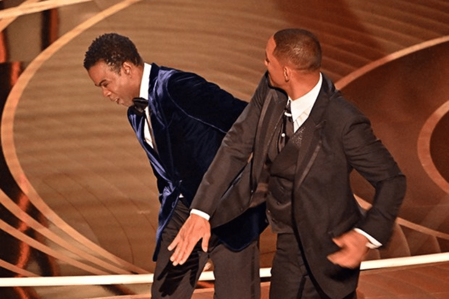 Two men in formal suits are on stage with a large, lighted background. One is gesturing with an outstretched arm towards the other, who is slightly bent forward.