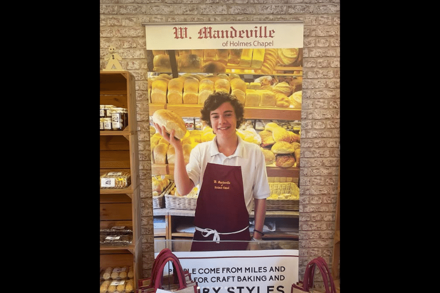 A bright, welcoming bakery display featuring a young person in a maroon apron, holding a loaf of bread. Shelves behind are lined with numerous golden-brown loaves, creating a warm, inviting atmosphere.
