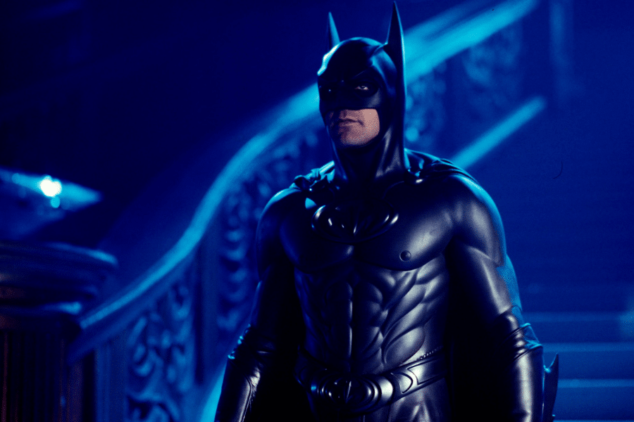 Batman stands solemnly in a dimly lit gothic mansion, wearing a detailed black suit with a bat emblem. Blue tones create a mysterious atmosphere.