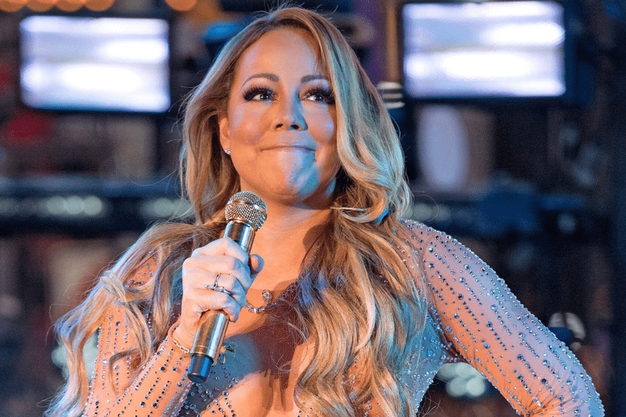 A person with long, wavy hair holds a microphone on stage, wearing a sparkly outfit. They appear thoughtful, with bright lights in the blurred background.
