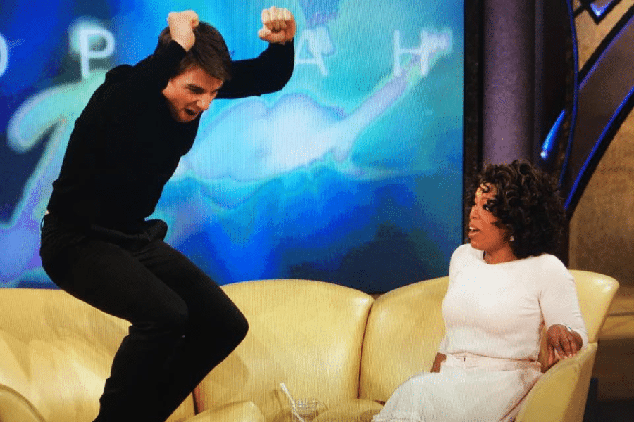 A man in black joyfully jumps on a yellow couch next to a seated woman in white. She appears surprised. A colorful, abstract backdrop is behind them.