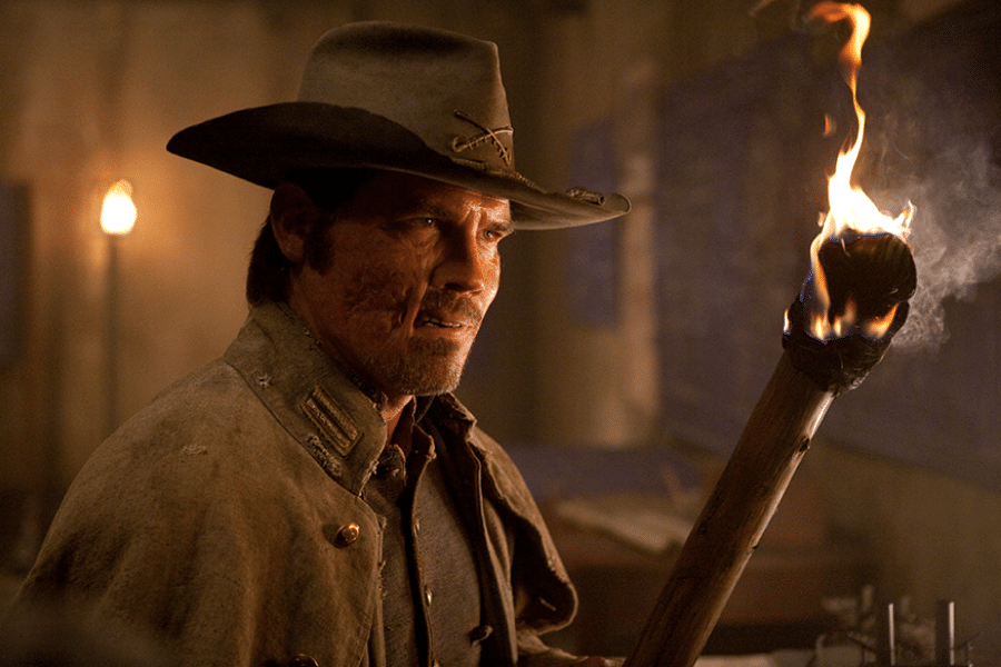 A rugged man wearing a cowboy hat and weathered coat holds a flaming torch in a dimly lit room. His expression is intense and focused.