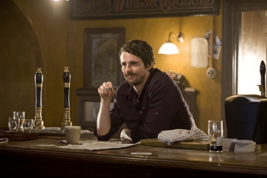 A man with short dark hair leans on a wooden bar adorned with glasses and a striped cloth. He smiles casually in a warmly lit pub setting.