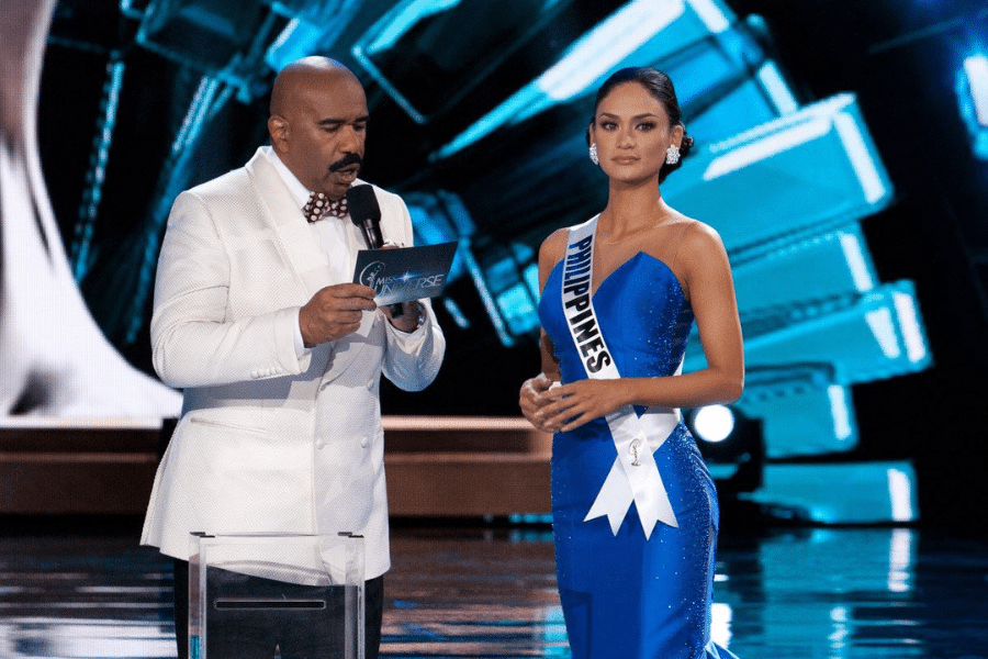 Host in a white suit, holding a microphone and card, stands next to a contestant in a blue gown and sash that reads "Philippines." The background is illuminated with blue and white lights, creating a formal and anticipatory atmosphere.