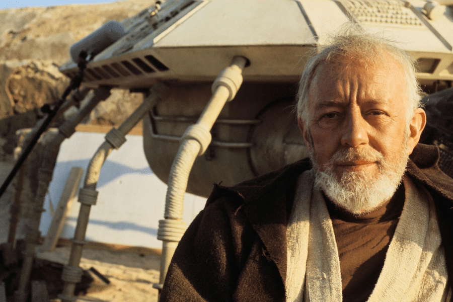 Elderly man with a beard and brown robe stands outdoors beside a futuristic vehicle on a desert landscape, conveying a sense of wisdom and resilience.