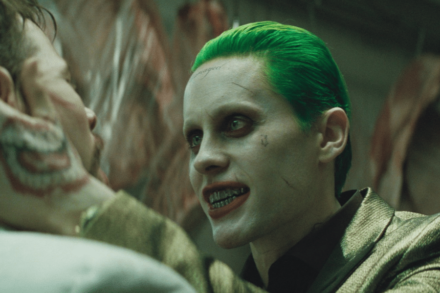 A man with slicked green hair, pale skin, and dark eye makeup menacingly faces another person. He wears a gold jacket and grills, conveying intensity and chaos.