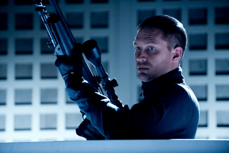 A man with short hair, wearing a dark jacket and gloves, holds a sniper rifle in a dimly lit, grid-patterned room. His expression is intense and focused.