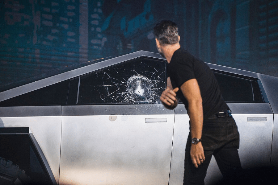 Man in black shirt looks at cracked window of futuristic, angular vehicle on stage. Scene conveys surprise and tension during a demonstration.