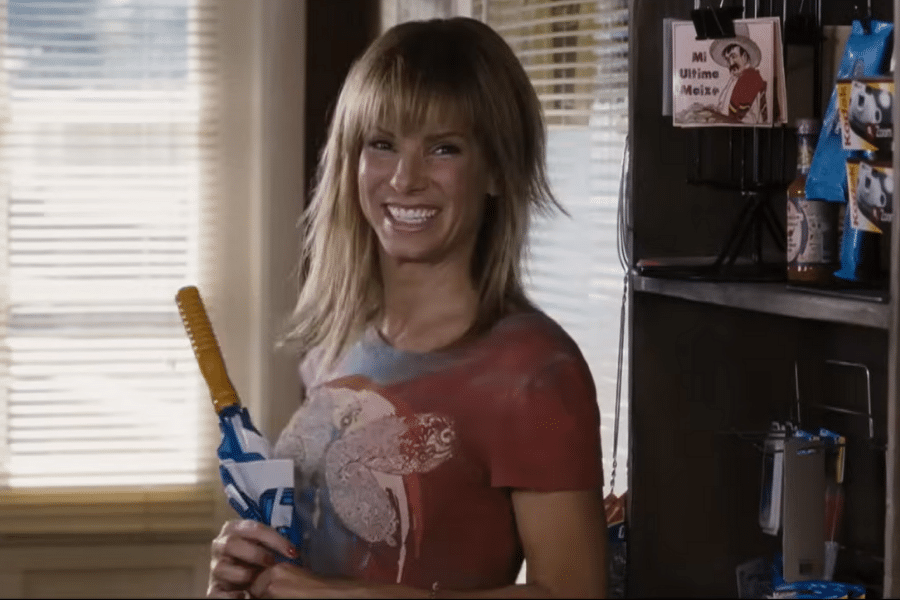 Smiling woman with layered hair holds a mop in a cozy room with sunlight through blinds. Shelves display snacks and a framed "Mi Último México" artwork.