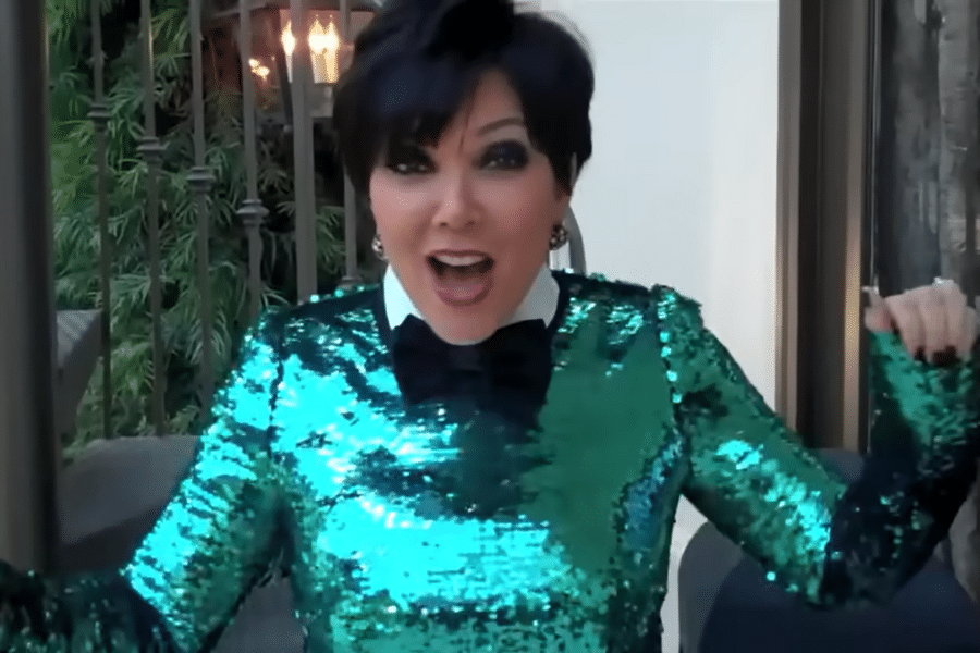 A person excitedly poses indoors, wearing a vibrant, sparkly green top and a black bowtie. The background features greenery and warm lighting.