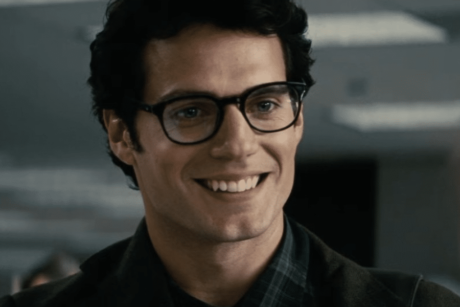 A smiling man with dark hair and glasses in an office setting. He wears a plaid shirt and blazer, conveying a friendly and approachable demeanor.