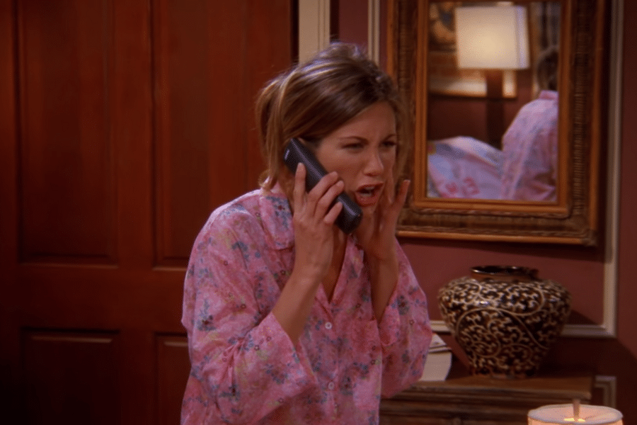 Woman in pink floral pajamas is on the phone, looking surprised or frustrated. Background shows a wooden door, ornate mirror, and decorative vase.