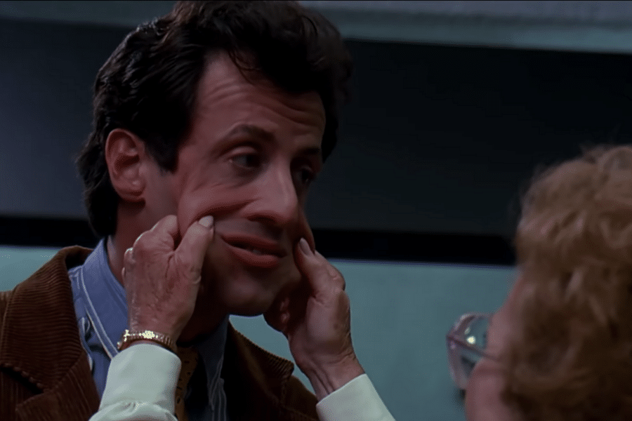A man with a playful, squished expression as his cheeks are pulled by a woman's hands. The setting appears to be a humorous, lighthearted moment.