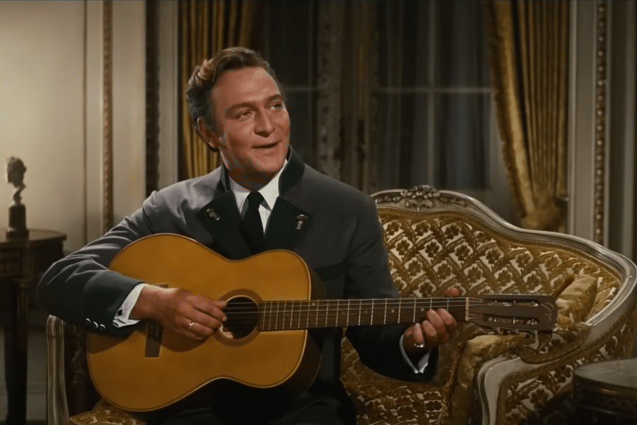 A man in a formal suit plays a guitar while seated on an ornate, patterned sofa. The room is elegant, with rich curtains and a warm, nostalgic tone.