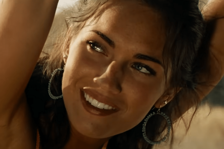 Smiling woman with hoop earrings looks to the side, arms raised overhead. Sunlit face, relaxed expression, earthy tones in the background.
