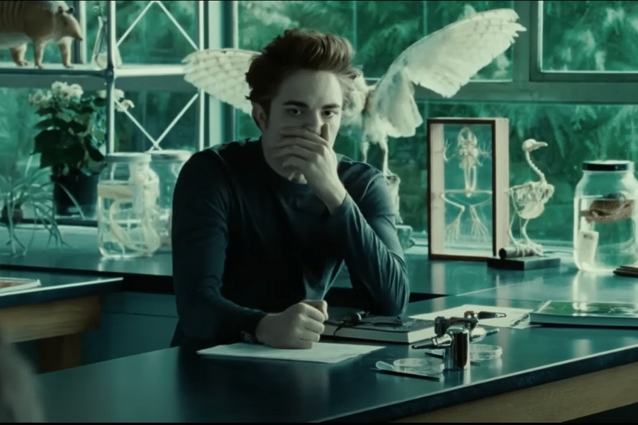 Young man with a serious expression sits at a science lab table, hand over mouth. Background includes jars and a bird model. Mysterious and intense mood.