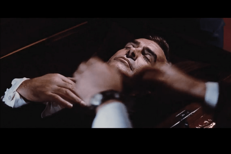 A man lies in a casket with eyes closed and hands clasped. The scene is dimly lit, conveying a solemn and tense mood, with blurred movement nearby.