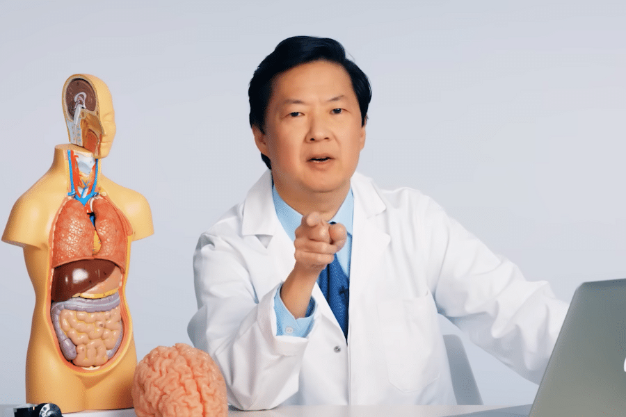 A person in a lab coat points at the camera while sitting next to a human anatomy model and a brain model. The setting is serious and educational.