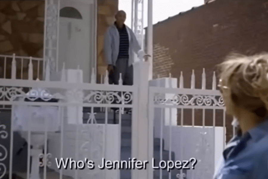 A person stands on a porch looking at another individual in the foreground, who is partially seen. A white fence separates them. The subtitle reads, "Who's Jennifer Lopez?" The scene is casual and inquisitive.