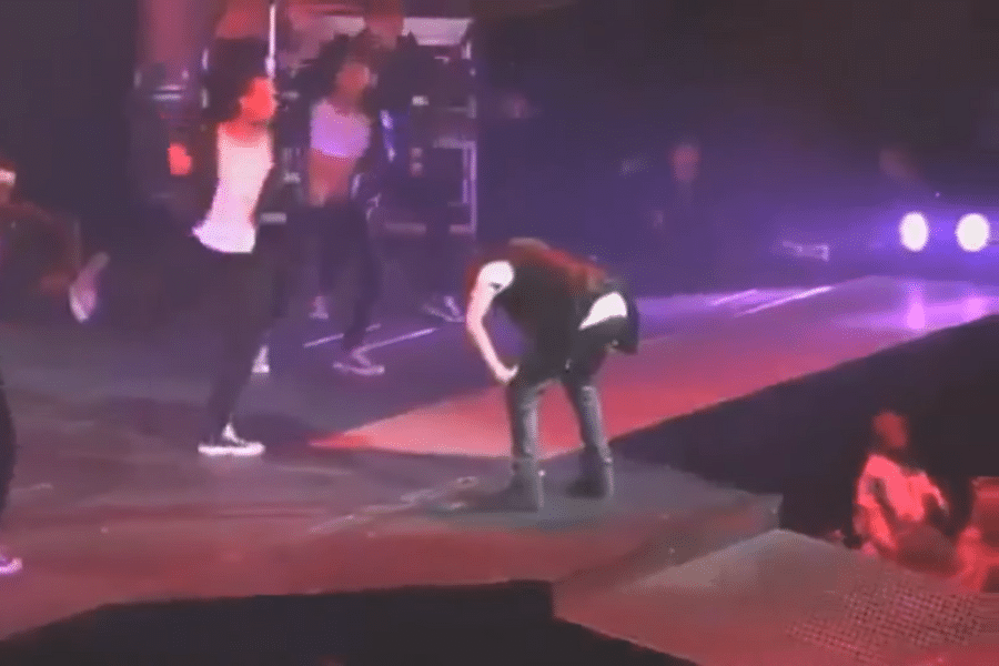 A performer bent over on stage puking, with backup dancers in the background. The stage is lit with purple lights, creating an energetic concert atmosphere.