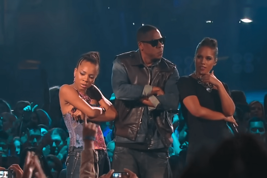 Three people stand on stage with arms crossed, exuding confidence. The atmosphere is lively, with a crowd in the background and blue lighting.