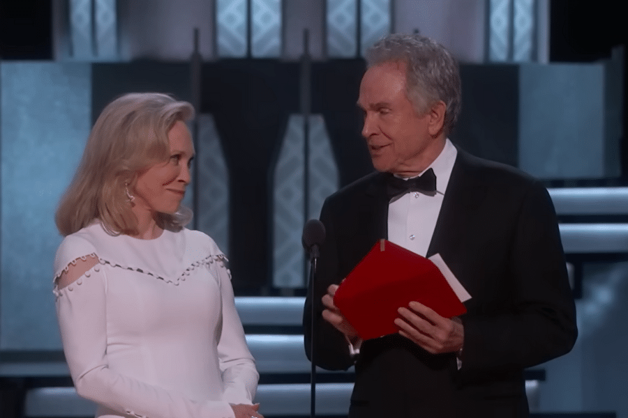 An elegantly dressed woman and man stand on stage, exchanging glances with slight smiles. The man holds a red envelope, suggesting an award event.