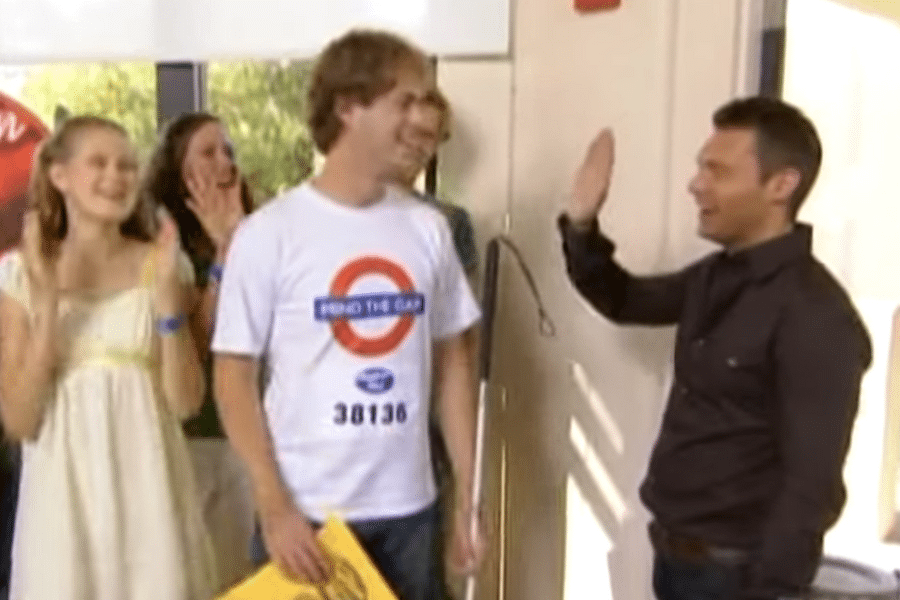 A cheerful group celebrates as a man in a "Mind the Gap" shirt holds a yellow envelope. Another man, smiling, raises his hand in a high five waiting for the other man. The mood is joyful.