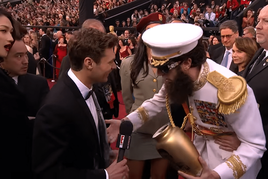 A man in a tuxedo interviews a bearded person in a decorated military outfit holding an urn on a crowded red carpet. The scene is lively and humorous.