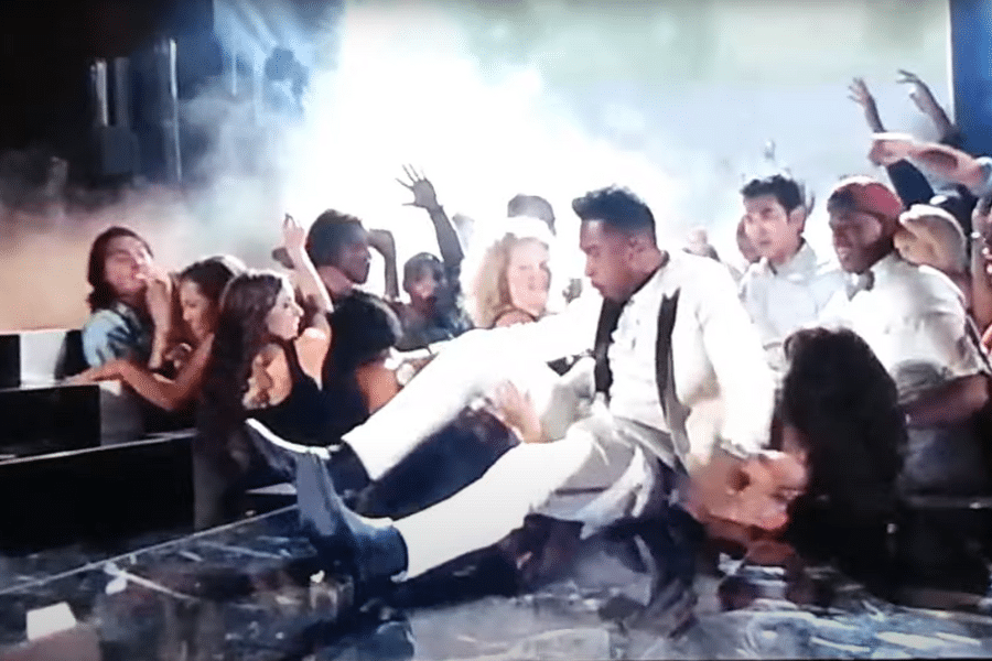 A performer in a white suit falls into a lively, cheering crowd with hands outstretched. Smoke and bright stage lights create an energetic atmosphere.