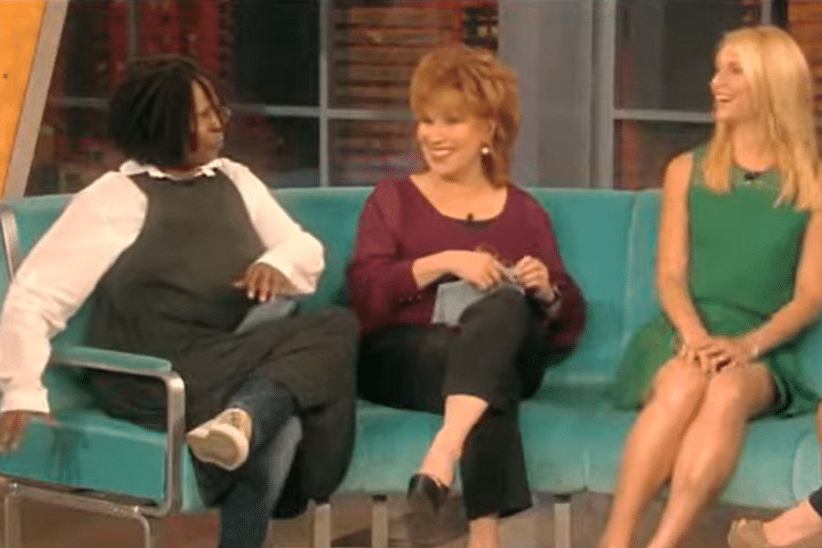 Three women are seated on a turquoise couch, smiling and engaged in conversation. The setting appears to be a talk show studio, creating a lively and friendly atmosphere.